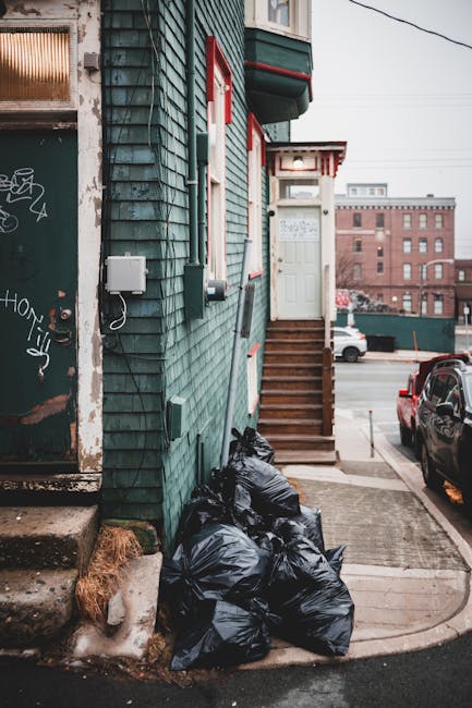 A narrow urban alleyway cluttered with discarded waste, featuring a large, partially full black and grey rubbish bag in the foreground, placed on a patchy, uneven asphalt surface. Behind it, an array 