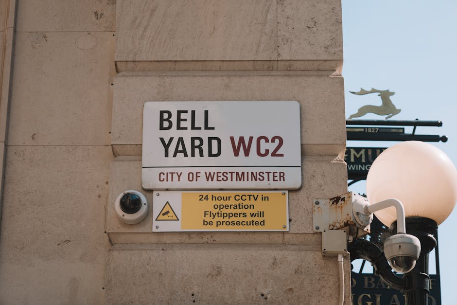 A close-up photograph of a beige stone building corner displaying a white rectangular sign with black and red text indicating 'BELL YARD WC2' and 'CITY OF WESTMINSTER.' Below, a smaller yellow sign wi